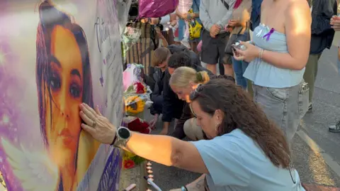 George King/BBC A woman crouching down in front of a large banner on which there is a photo of Courtney Mitchell's face. To her right there are other men and women crouching down lighting candles.