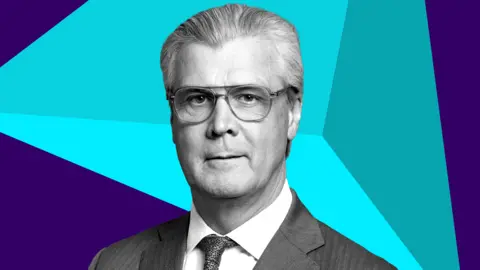 Malcolm Offord, who has grey hair swept back and glasses, in a down the lens shot. He is wearing a business suit, with a blue digitised graphic behind him. 