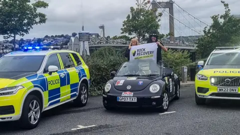 Devon and Cornwall Police A man and woman stand on the front seats of an open top Volkswagen Beetle, holding a sign saying Rock 2 Recovery. The car is flanked by two police cars and the Tamar bridge is in the background.