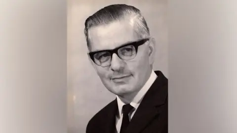 Jane Martin Fred Martin pictured as an older man, wearing a pair of 1960s style glasses with thick frames on top and no frame below. His hair is slicked to one side and he is looking just off camera with a gentle smile. It appears to be in black and white. He is wearing a dark suit and tie and white shirt.