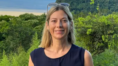 Family handout A woman with short blonde hair wearing sunglasses. She is smiling at the camera and is wearing a blue top. Behind her are green trees.
