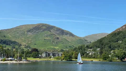 Susan View of a lake with a country house type hotel on grounds leading down to it. A small white sailed boat is on the water and to the left other small sailing boats are moored in an inlet. A mountain rises in the background under blue skies.