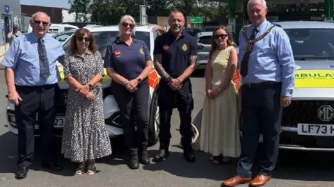 Debbie is pictured with members of the CFR team and a mayor and they are standing in front of two vehicles. Debbie is in a patterned sundress and Ellie is wearing a yellow dress. A male and female responder are wearing a navy uniform two other men, on the far left and right, are wearing a navy shirt and trousers. The man on the right is wearing mayoral chains over his shirt




Tim Bridgland-Taylor (Hereford Freemasons), Debbie Roscoe(Arrive Alive Founder), Fran Thomas (community first responder), Daryl Szebesta (community first responder) Ellie Roscoe (Debbie’s daughter), Stuart Jones (Hereford Freemasons)
