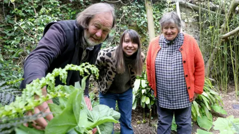 Rick Matthews A man and two women is stood in a row. The man on the left has grey hair and beard, is wearing a black fleece, and is leaned down, touching a green plant. The woman in the middle has long brown hair and is wearing blue jeans and a brown leopard print fleece. The woman on the right is wearing grey jeans, a check shirt and an orange cardigan. She has short grey hair. They are all stood in woodland with shrubbery around them and are smiling as they look at the camera.