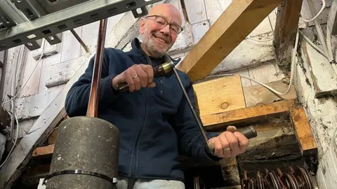 Graham Tebbs inside the clock tower holding a long winding handle. Around him are wooden beams. Mr Tebbs is wearing a navy blue zip-up fleece and has white receding hair, a white beard and glasses. He is smiling.