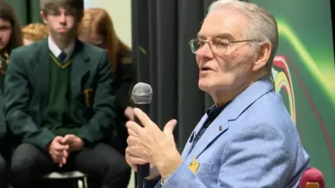 Tomi Reichental is wearing a light blue shirt. He is wearing a black shirt and is holding a microphone. Some school children are visible in the background. 