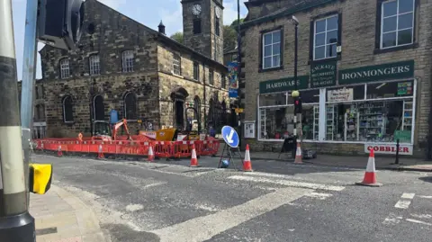 Hayley Coyle/BBC A road in Holmfirth with road words, traffic cones and a mini digger, in front of a row of shops.