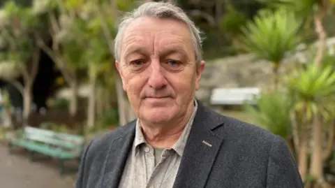 Anthony Slaughter, standing in a public park looking at the camera. There are palm trees and a park bench in the background. He is wearing a light grey shirt, open at the neck, and a dark grey jacket. He has short, grey hair.