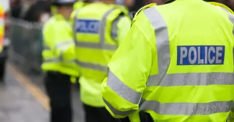 Three Police Officers standing with yellow hi-vis jackets on which read 'Police'.