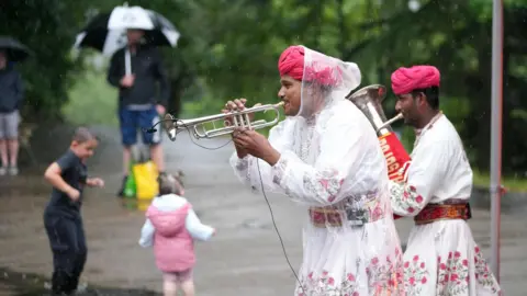 Durham County Council Two men play a trumpet and trombone in a park as it is raining. Two kids can be seen playing in the background.