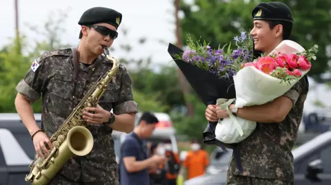 Getty Images RM, Kim Namjoon is wearing his military uniform and sunglasses, he is playing the saxophone. V, Kim Tae-hyung, is looking at RM, also clad in military uniform, holding two bouquets of purple and pink flowers.  
