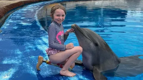 Natalia grins as she kneels down on her left prosthetic leg as she strokes a dolphin by the side of a pool. 