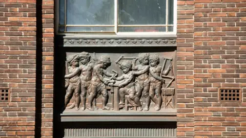 Historic England A bronze  plaque set into the wall of the building, showing six child figure working on parts of a bicycle