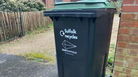 Vikki Irwin/BBC A large black wheelie bin pictured outside a driveway entrance. It is a black bin with a green lid. On the bin it says "Suffolk recycles" and "East Suffolk Council". 