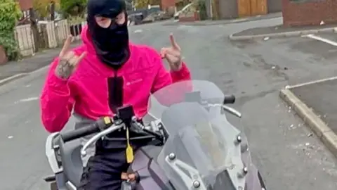 Teen poses on a bike wearing a balaclava making hand gestures