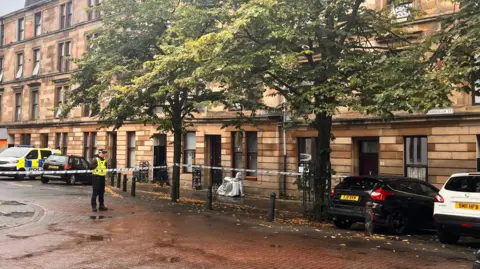 A long street of blonde sandstone tenements with some cars parked in front of them. Part of the street is corned off with Police tape, and a police officer is visible behind it.