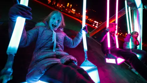 Andre Pattenden Three people swing on illuminated swings which have been placed in Bristol as part of the Bristol Light Festival. The person closest to the camera, a young woman, is smiling with her mouth open. 