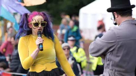 A woman with two hair bunches that are dyed pink and purple is singing into a microphone. She is wearing black and yellow stripy sunglasses and has black and orange honeycomb style face paint. She is wearing a yellow top and belt