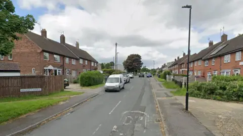 A road with houses on either side. Cars parked on both sides of the road.