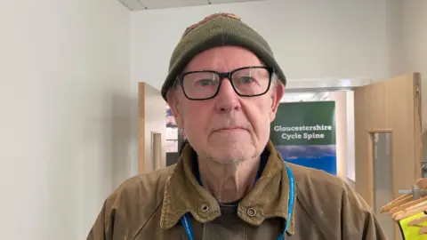 A man is standing in front of an open door way. He is wearing a green hat, glasses and a brown jacket. 