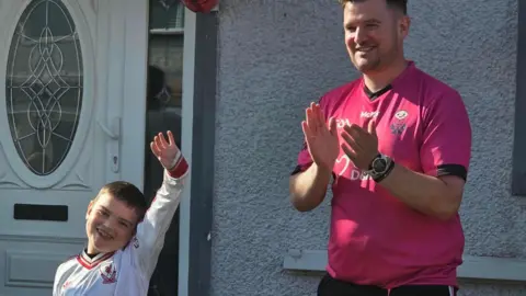 Dáithí is holding his hand up in the air. His dad Máirtín is clapping.