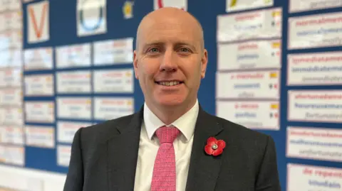 BBC man with bald head and hair tightly shaved at sides wearing grey suit and pink tie, wearing red armistice poppy pin, stands in front of wall of school work