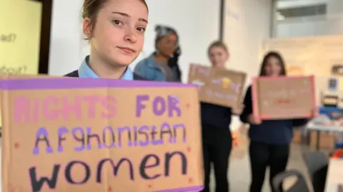 A school girl holding a hand painted placard which reads Rights for Afghanistan Women. Two other pupils stand in the background.