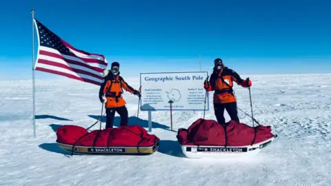 Jordan Wylie Jordan Wylie and another member of the team stand on a large flat expanse of ice under a blue sky at a sign that says "Geographic South Pole". There is an American flag to the left and the men are wearing black and orange coats and masks and have sledges of equipment covered with red tarpaulins