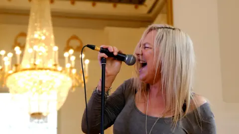 Kady Braine/BBC A woman with long blonde hair wearing a grey long sleeved top singing passionately into a microphone with a chandelier in the background