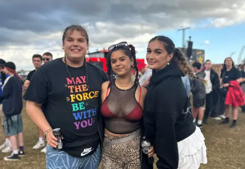 A man and two women stand in a field in front of the main stage at Reading Festival. They're holding drinks and smiling.