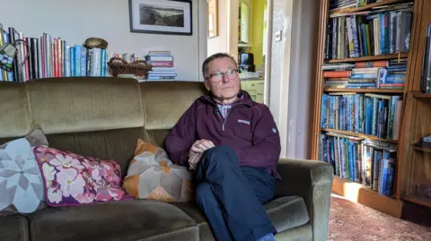 Andrew, a man with short gray hair and glasses stares away from the camera and into the middle distance. He is wearing a burgundy zipped jumper and sitting on a brown sofa in his living room.