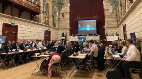 A room with lots of people sat at desks and chairs. 