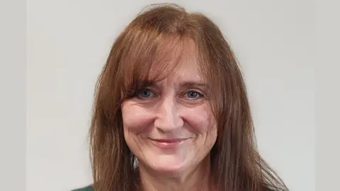 A woman with long brown hair stands against a grey background and smiles at the camera in a corporate council head-shot.