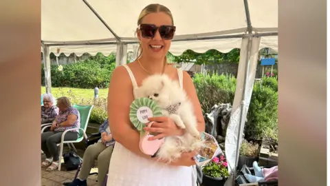 Natalie Crampton A woman in a white dress holding a white dog with a green rosette which says "Puppy love"