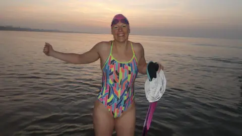 Elizabeth Mills Elizabeth Mills in a multi-coloured swimming costume and a pink swimming cap emerging from the sea.