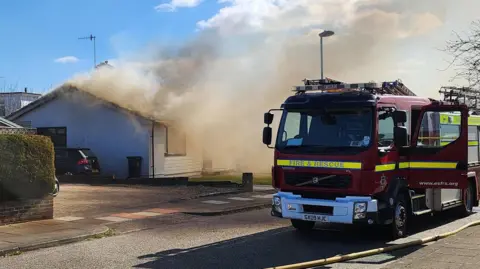 Eddie Mitchell Smoke pours from a bungalow on a sunny day. 