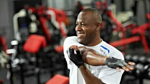 Getty Images A man performing a shoulder stretch warm up exercise before workout, smiling in gym.