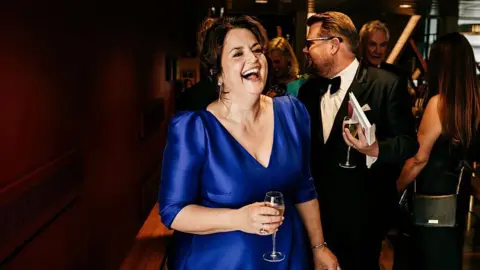 Getty Images Ruth Jones in a blue dress has a giggle as she holds a glass of champagne. Her co-star and co-writer James Corden is just behind her.