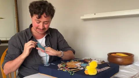 Florian Barrows sitting at a desk, carefully manipulating blue lace over a dark blue pillow packed with wooden bobbins and a small yellow rubber duck.