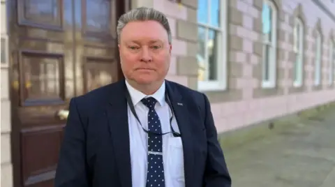 Rob Ward is wearing a black blazer, white shirt and a polka dot tie. 
