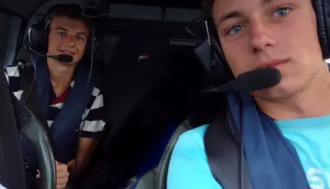 Ben West Ben and Sam are in a vehicle with headseats and a mic. Ben is wearing a blue t-shirt and Sam is wearing a stripy t-shirt, they are both smiling at the camera 