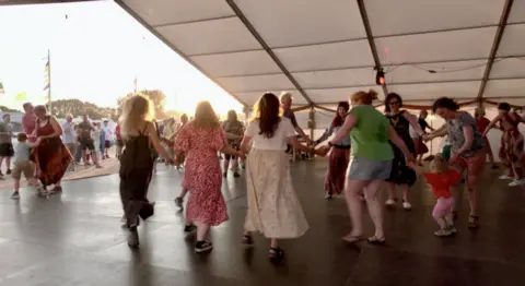 A group of people holding hands in a circle on a stage at an outdoor festival
