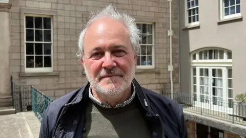 A white man with white hair standing in front of a grey building with green railings and white pane windows. He is wearing a dark jumper and open jacket with the collar of a checked shirt underneath.