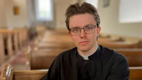 The Reverend Dr Edmund Smith is sat on a wooden pew, with rows of wooden pews behind him. He is looking directly at the camera and is wearing glasses.