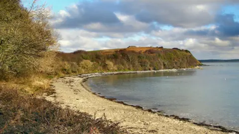 Penrhos Coastal Park a two hundred acre park on the outskirts of Holyhead on the coast. The photo shows bushes above a beach that stretches around bay to a forest and field covered point. 