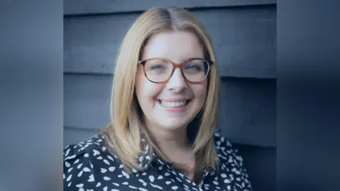 Head-and-shoulder shot of Hannah in a blue and white blouse. She has long blonde hair and glasses. She is smiling.