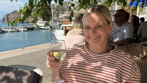 Rachel Bevan Rachel Bevan pictured holding a glass of white wine in her left hand. Rachel wears a red and white striped t-shirt, has blonde hair pulled back into ponytail, blue eyes and smiles with her teeth. A harbour can be seen behind her on the left. Tables filled with people at a restaurant can be seen behind her, sitting below trees in the shade. 
