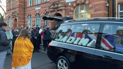 A black hearse with a Union Jack covered coffin in the back. A semi circle of people stand around the back of it.