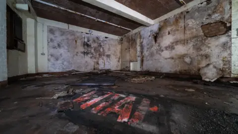 Cadw The indside of an underground bunker in disrepair. The room is mostly empty, with dirty walls and debris on the ground.