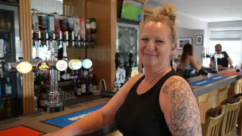 Shaun Whitmore/BBC A smiling woman sits at a pub's wooden bar, her right arm resting on the bar and her body and face turned towards the camera. She is wearing a black vest top. Her hair is blonde and tied back. Behind the bar are chrome beer taps and spirits bottles on optics. 